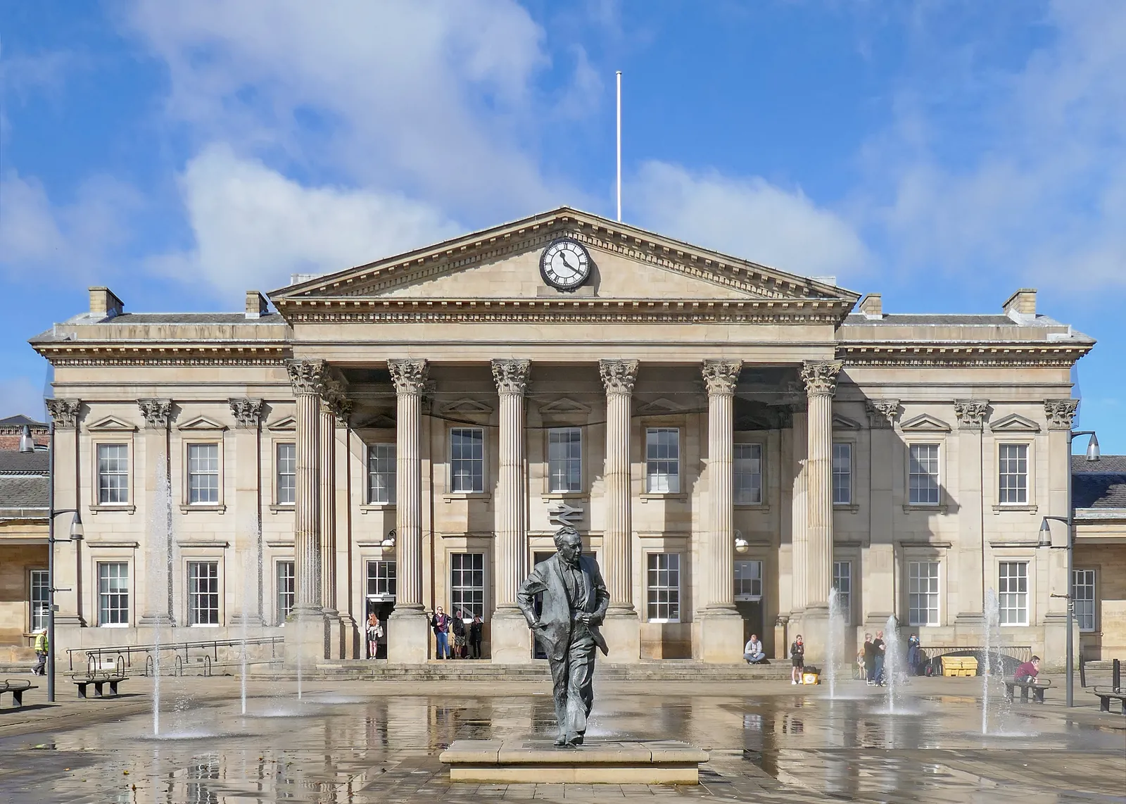 The columned front of Huddersfield railway station on St George's Square, with fountains and the statue of Harold Wilson